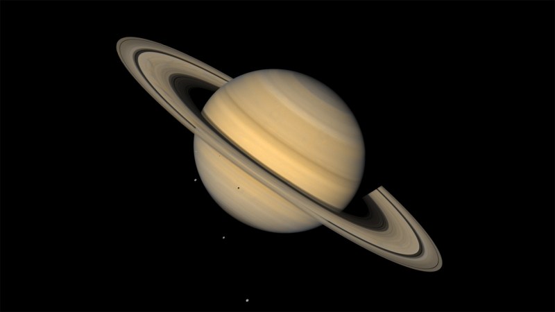 This image shows Saturn, ringed and in tones of tan, against an inky background. Three tiny light spots in the background and one dot against the lower half of the planet are four of Saturn